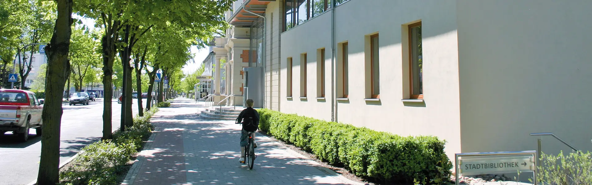 Eine Person fährt mit dem Fahrrad auf einem breiten Gehweg an einem sonnigen Tag. Links vom Gehweg verläuft eine von Bäumen gesäumte Straße mit parkenden und fahrenden Autos. Rechts befindet sich ein modernes, helles Gebäude, entlang dessen Fassade eine Reihe schmaler Fenster und eine ordentlich geschnittene Hecke zu sehen sind. Ein Schild im Vordergrund rechts weist mit der Aufschrift „STADTBIBLIOTHEK“ und einem Pfeil den Weg zur Stadtbibliothek.