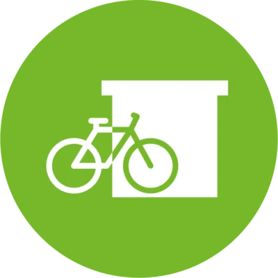 Weißes Piktogramm eines Fahrrads vor einem geschlossenen Abstellraum auf grünem Kreis – symbolisiert einen sicheren Fahrradstellplatz.